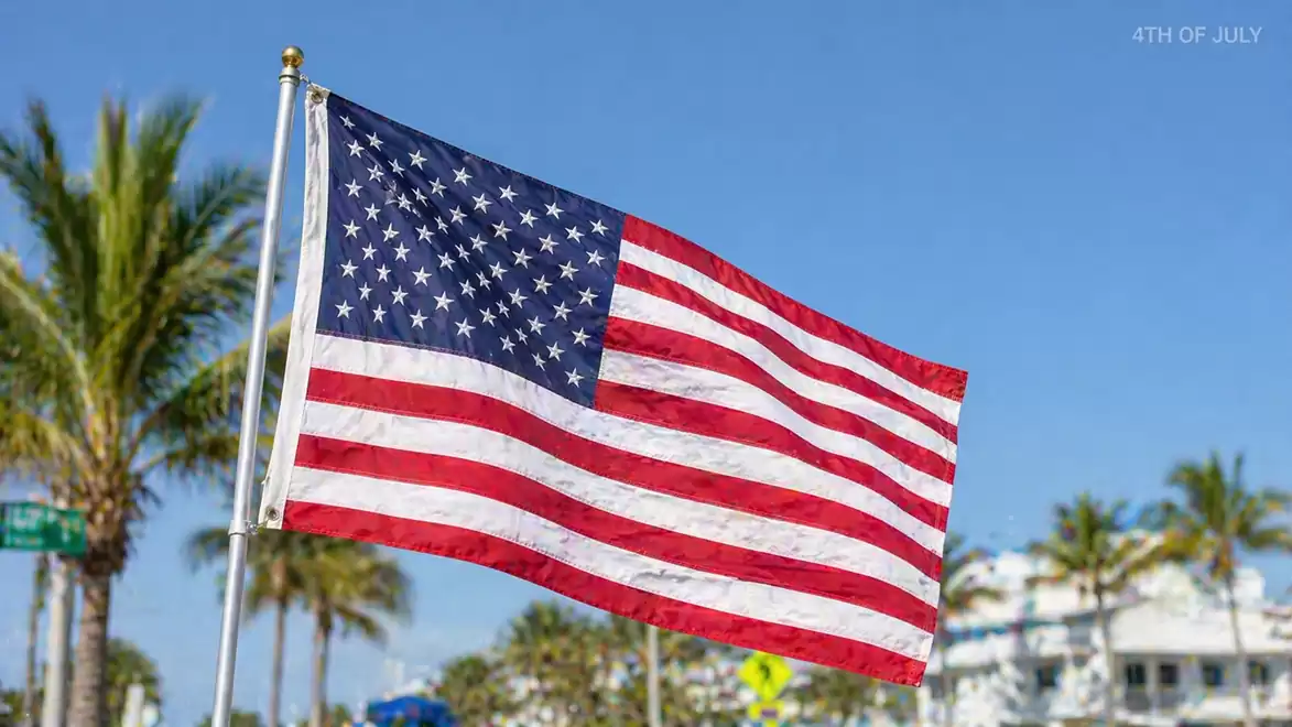 The American flag waves in the wind against a background of palm trees and a clear sky.