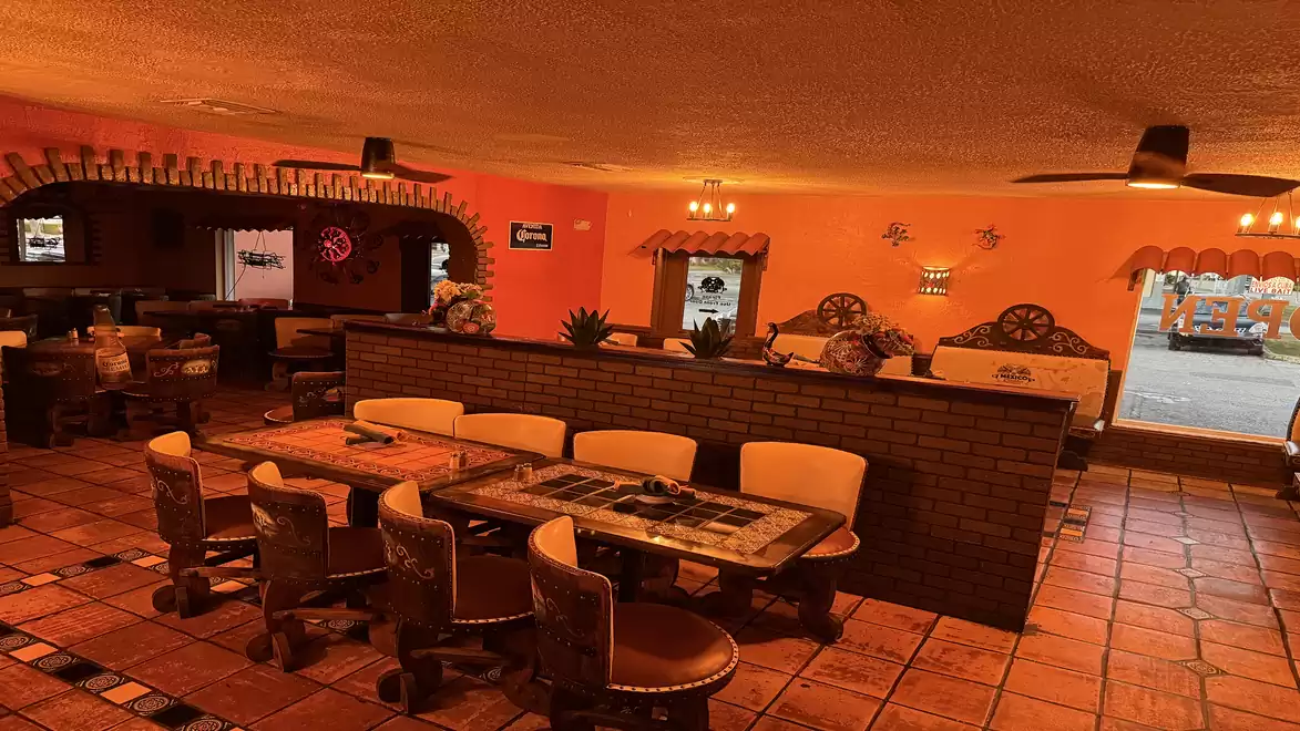 Interior view of a cozy, rustic restaurant with warm tones, wooden tables, and upholstered chairs. Decorations and a bar are visible in the background.