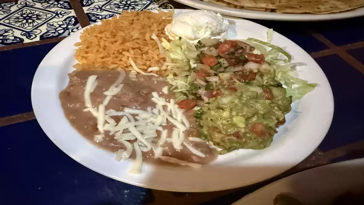 A plate of Mexican food, including rice, beans with cheese, salad, and guacamole. The background features a blue tile pattern.