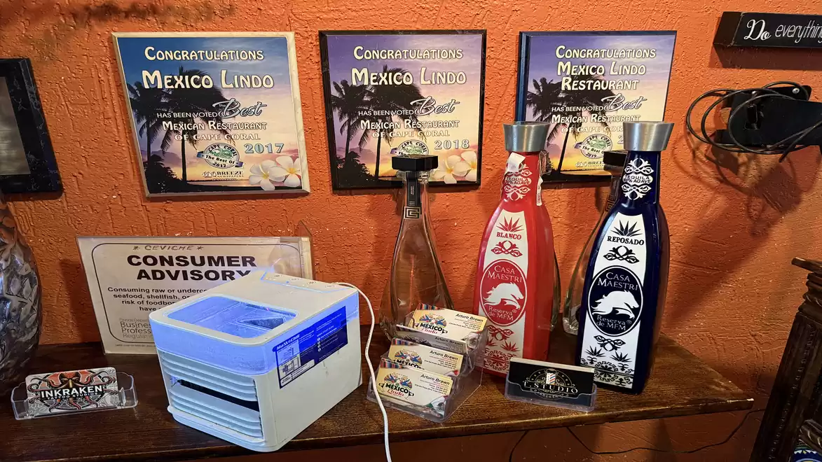 A table with awards from the 'Mexico Lindo' restaurant, decorated with colorful tequila bottles and gadgets. In the background, framed certificates hang on an orange wall.
