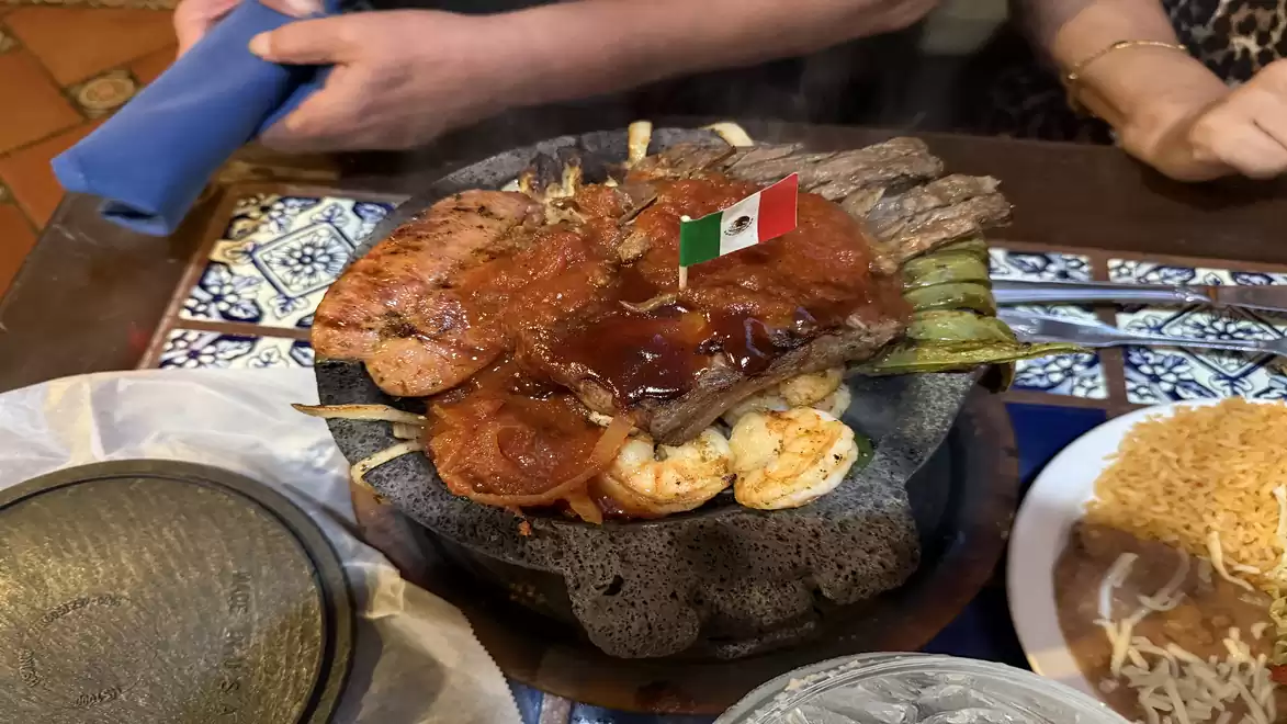 A mixed grill platter with various meats, a Mexican flag decoration, and side dishes on a table in a restaurant.