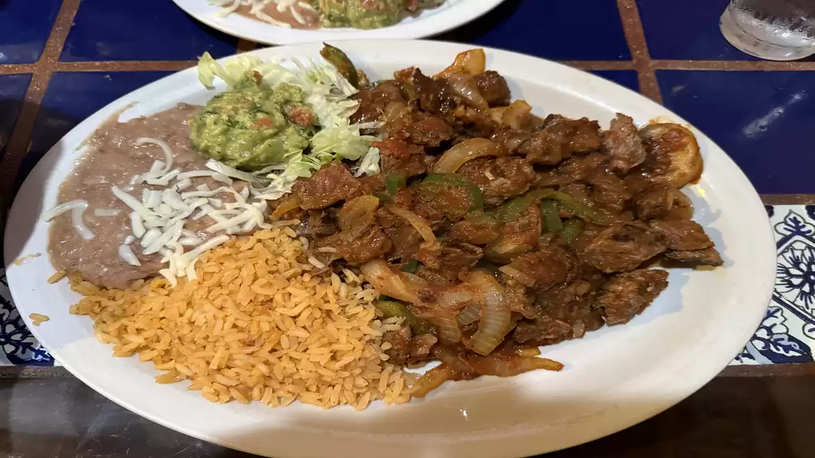 A plate with Mexican food consisting of fried meat, rice, beans, guacamole, and salad, served on a blue and white tiled table.