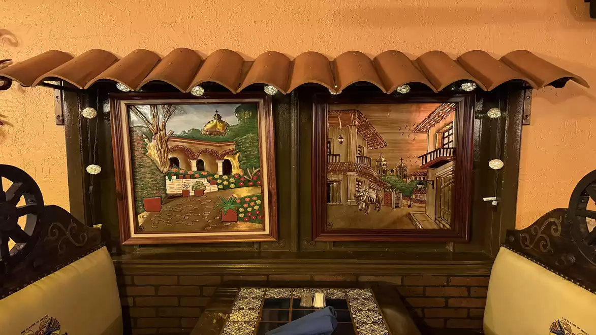 A cozy restaurant interior featuring two framed paintings on a brick wall. On the left is a landscape with trees and a domed building. On the right is a street scene with old balconies and lanterns. A decorative row of roof tiles above the images adds rus
