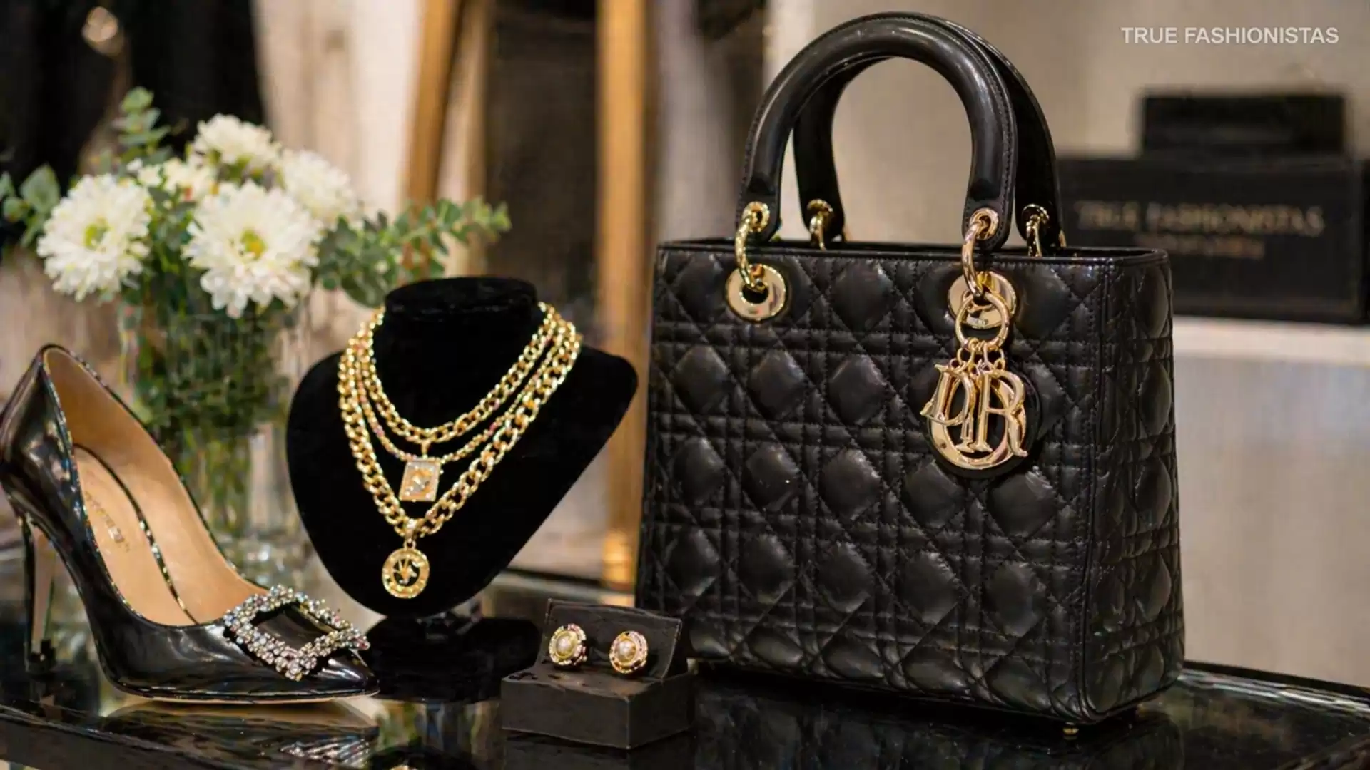 Luxurious display featuring a black designer handbag, elegant jewelry, stylish shoes, and flowers in the background.
