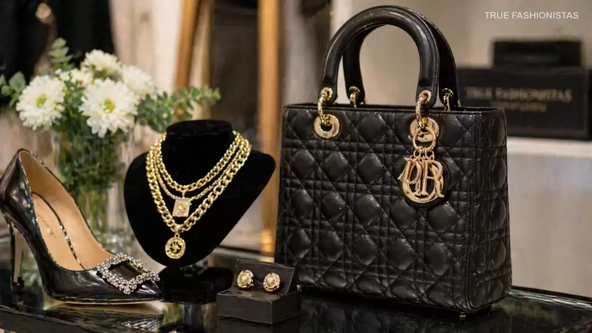 Luxurious display featuring a black designer handbag, elegant jewelry, stylish shoes, and flowers in the background.