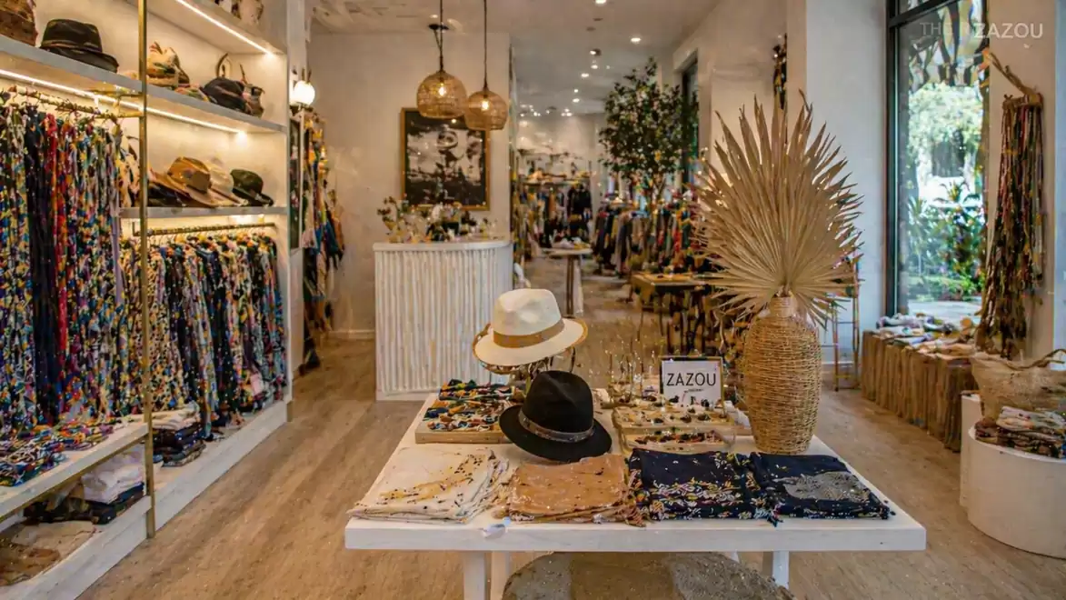 A stylish fashion store featuring laid-out scarves, hats, and decorations on a table, surrounded by shelves full of colorful accessories and modern lighting.