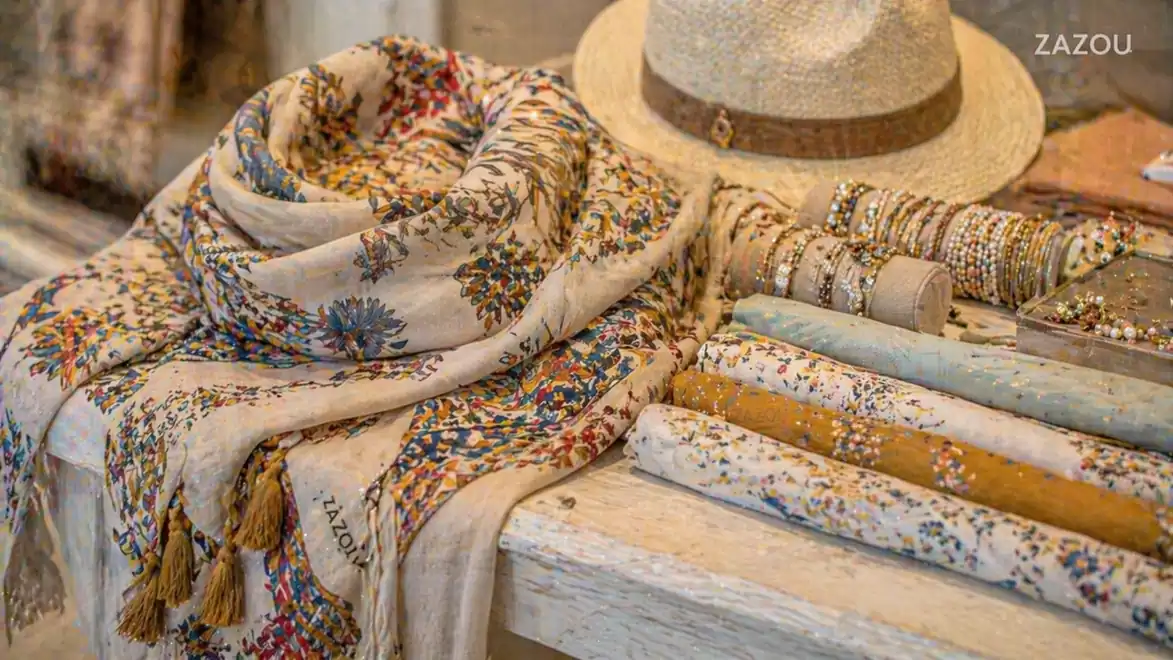 An elegantly arranged summer scarf and straw hat on a table with golden jewelry and colorful fabric rolls. Perfect for summer fashion.