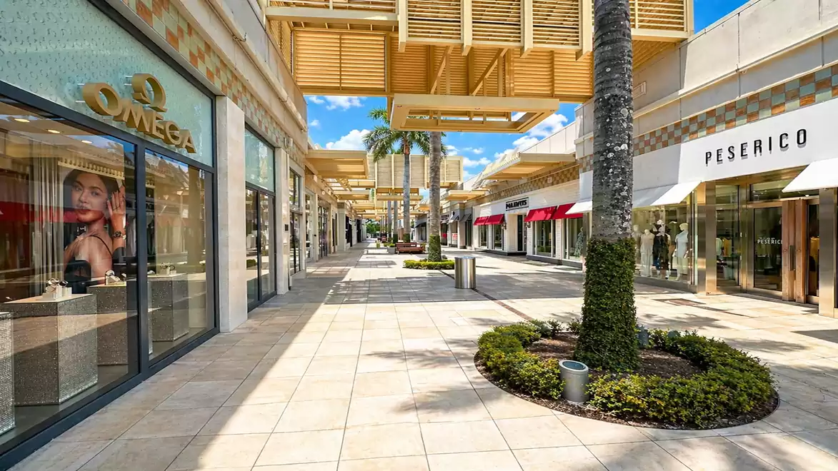 Shopping mall with luxury stores, including Omega and Peserico, set along a modern outdoor walkway with palm trees and blue sky.