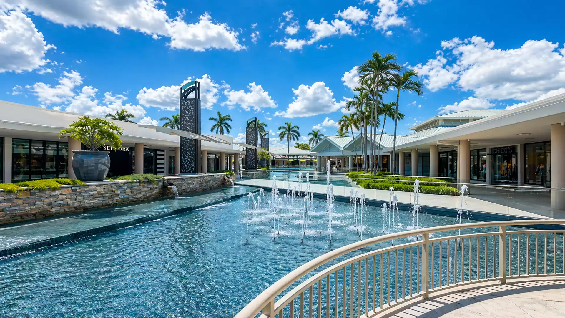 A modern shopping center with a large water fountain in the center, surrounded by palm trees and modern buildings under a clear blue sky.