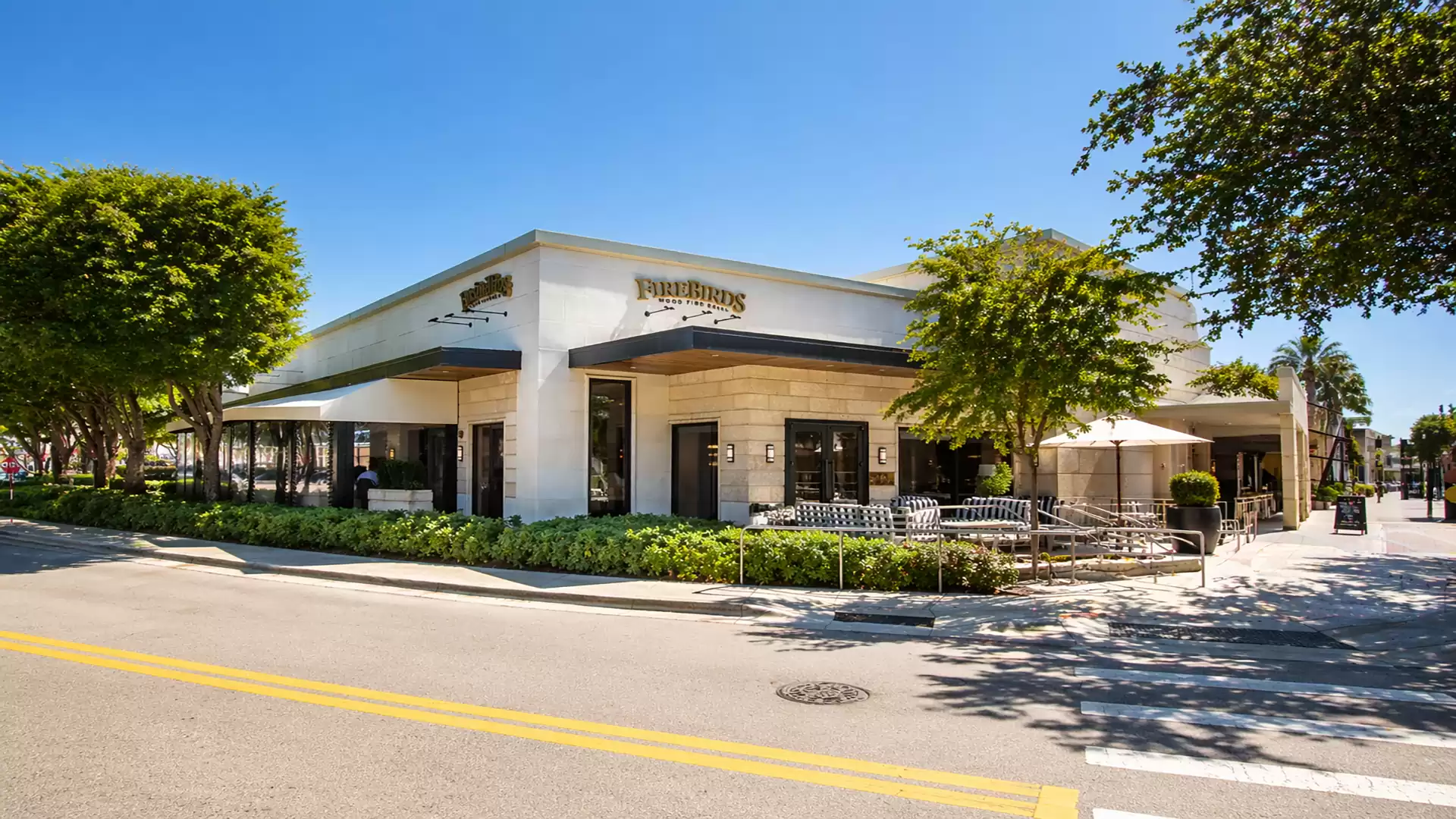 Exterior of a corner cafe under blue skies, surrounded by trees and sunny weather.