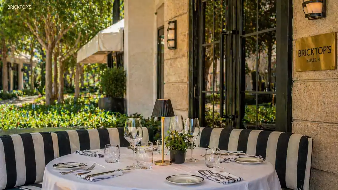 Elegant outdoor restaurant table with white tablecloth and striped seat cushions in a sunny garden setting.