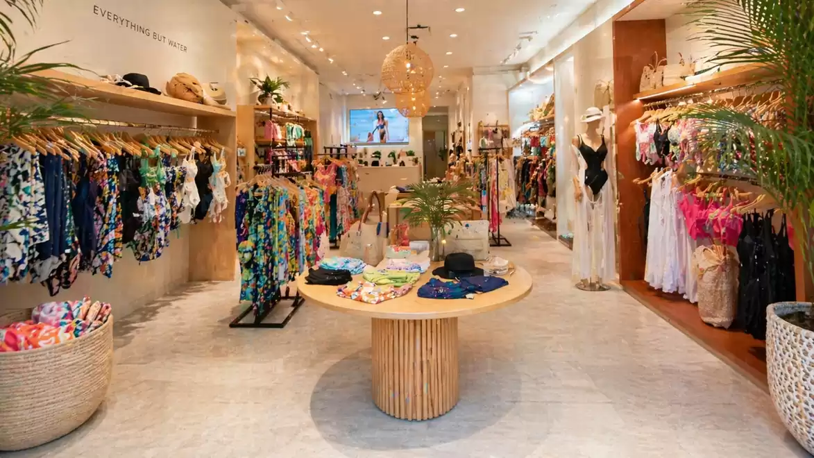 A stylishly arranged clothing store with summer clothes and accessories displayed on wooden shelves and a round table.