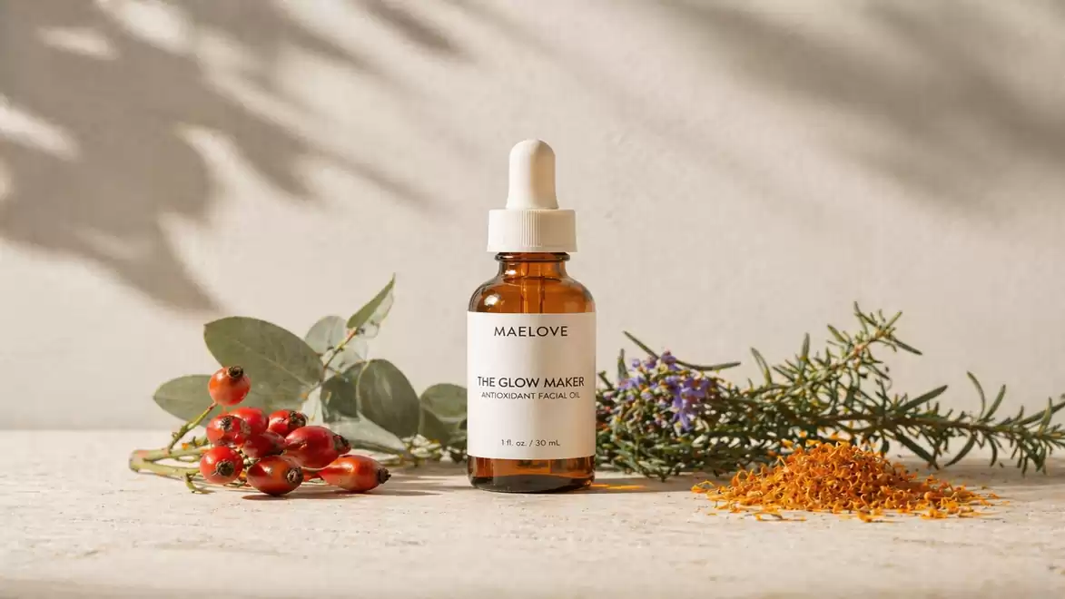 A dropper bottle labeled 'Maelove The Glow Maker', surrounded by herbs and berries on a light surface.