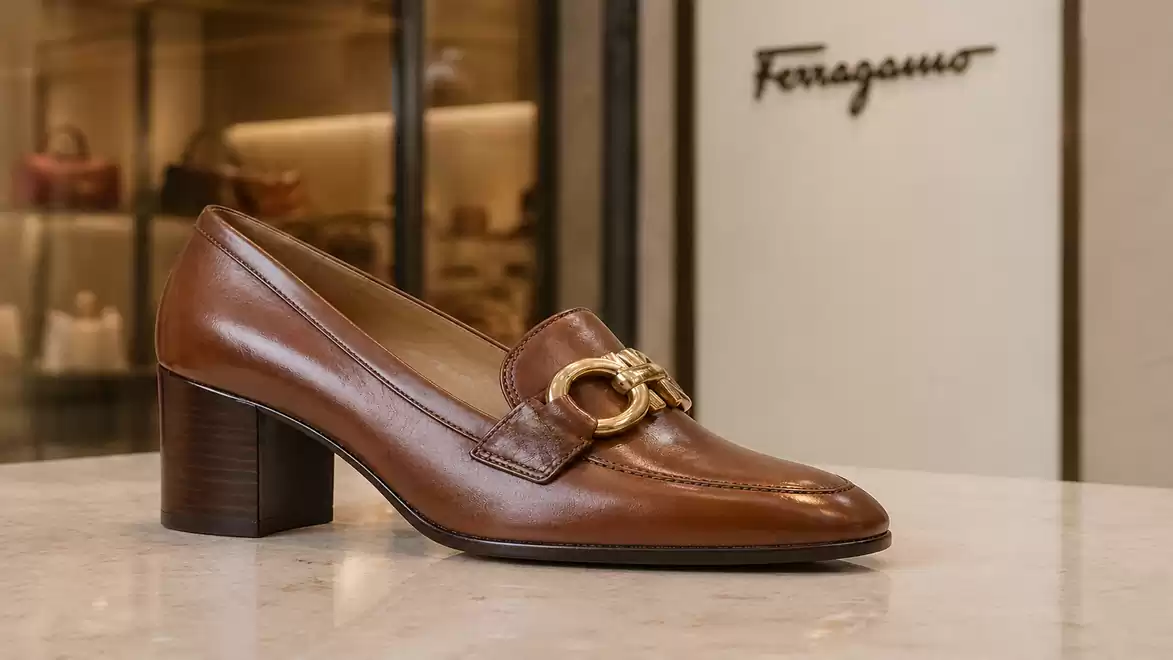 Brown leather shoe with heel and gold buckle in front of a Ferragamo boutique.