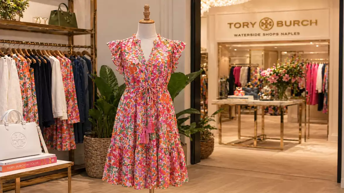 An elegant floral dress on a mannequin in a stylish clothing store with tables and shelves full of clothes and accessories.