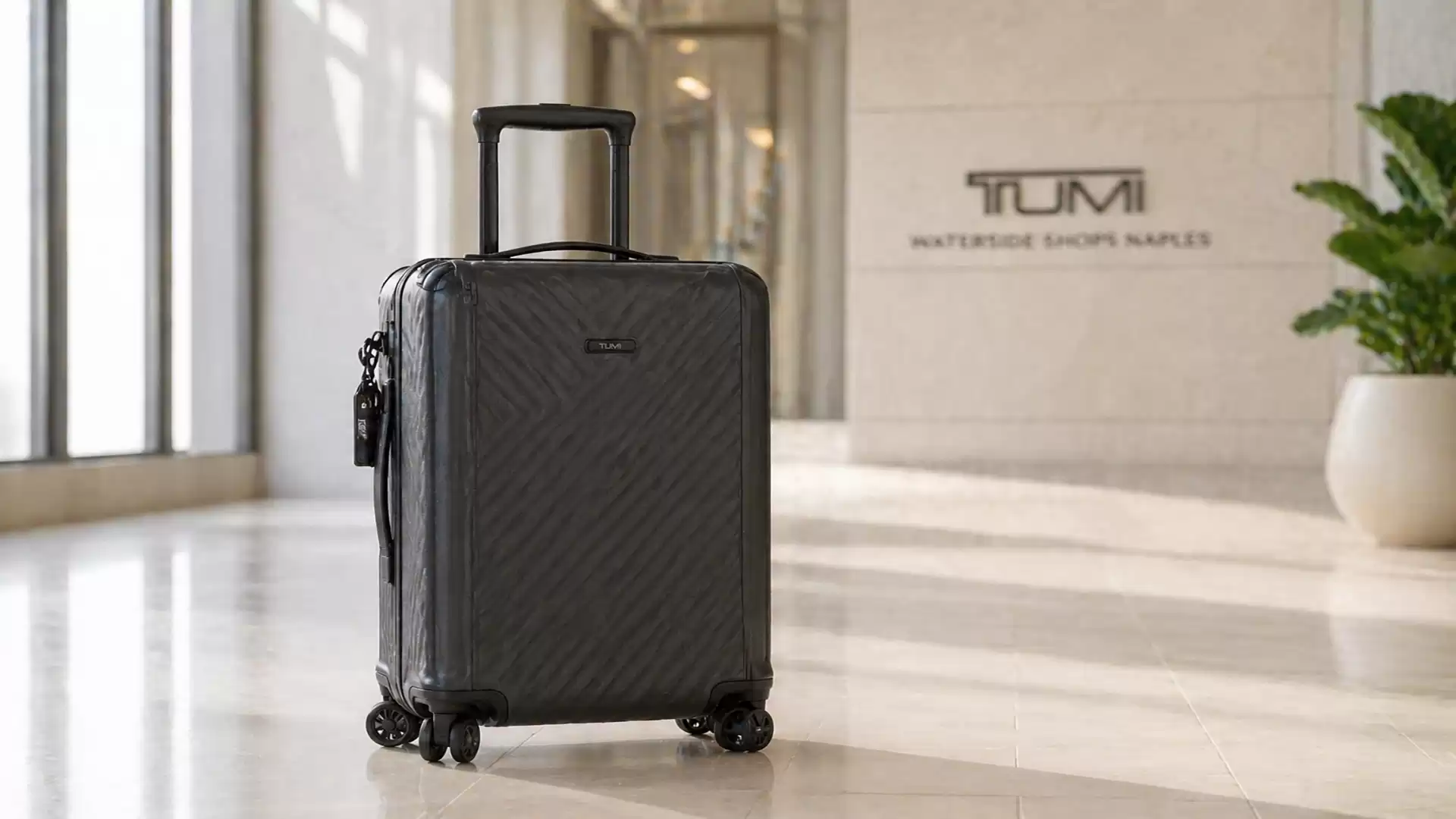 A sleek, black trolley suitcase stands in a modern building with a Tumi logo in the background.