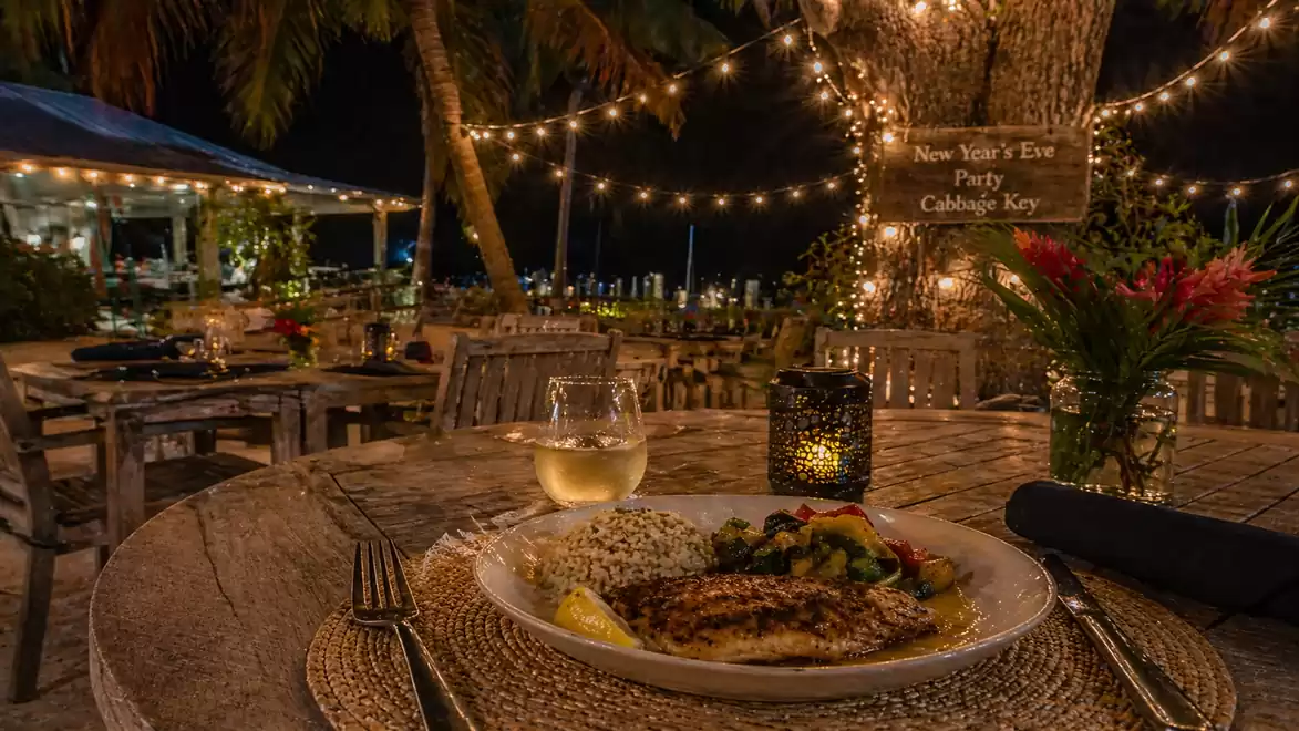 A beautifully lit outdoor restaurant area at night, featuring a decorative table with a delicious dinner plate under a tree adorned with string lights.