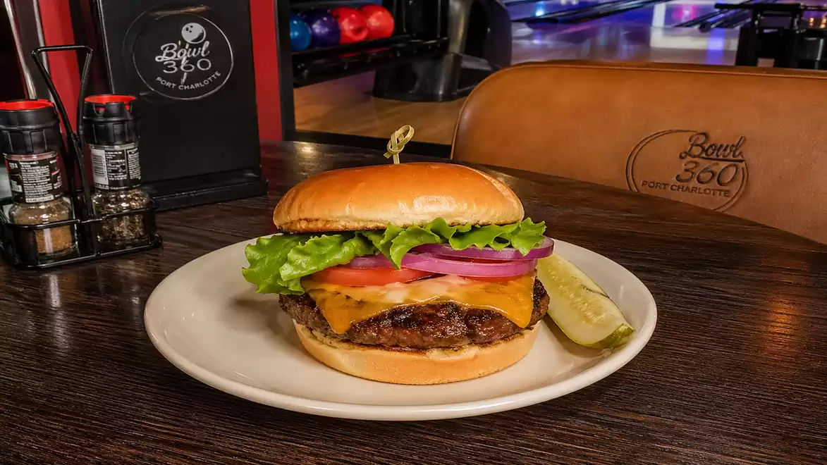 A delicious cheeseburger with lettuce, tomato, and onion on a plate, garnished with a pickle slice, with a bowling alley in the background.