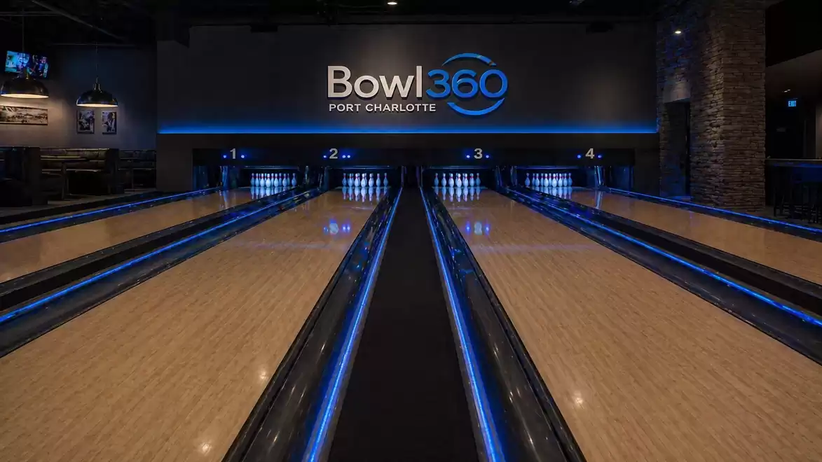 Interior view of a modern bowling alley, illuminated with blue lights. The sign 'Bowl 360 Port Charlotte' is visible in the background, surrounded by multiple lanes.