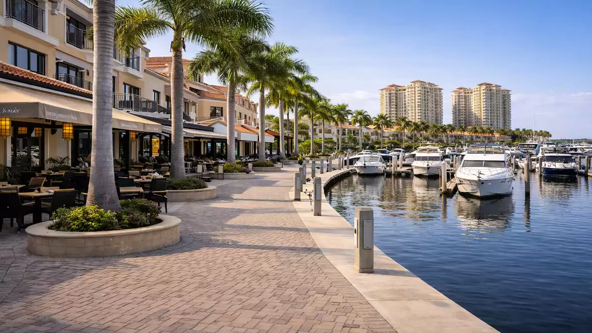A picturesque marina with modern residential buildings in the background. Elegant boats are docked at the pier, with palm trees lining the paved promenade. Inviting cafes and restaurants offer seating with a view of the water.