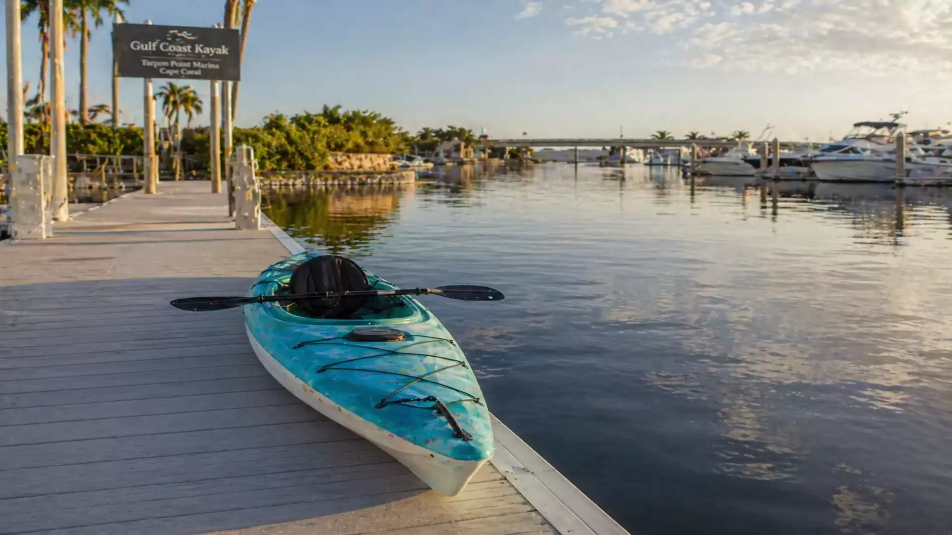 A blue kayak lies on a wooden dock in a marina with several yachts. The sky is partly cloudy and the sun is shining.