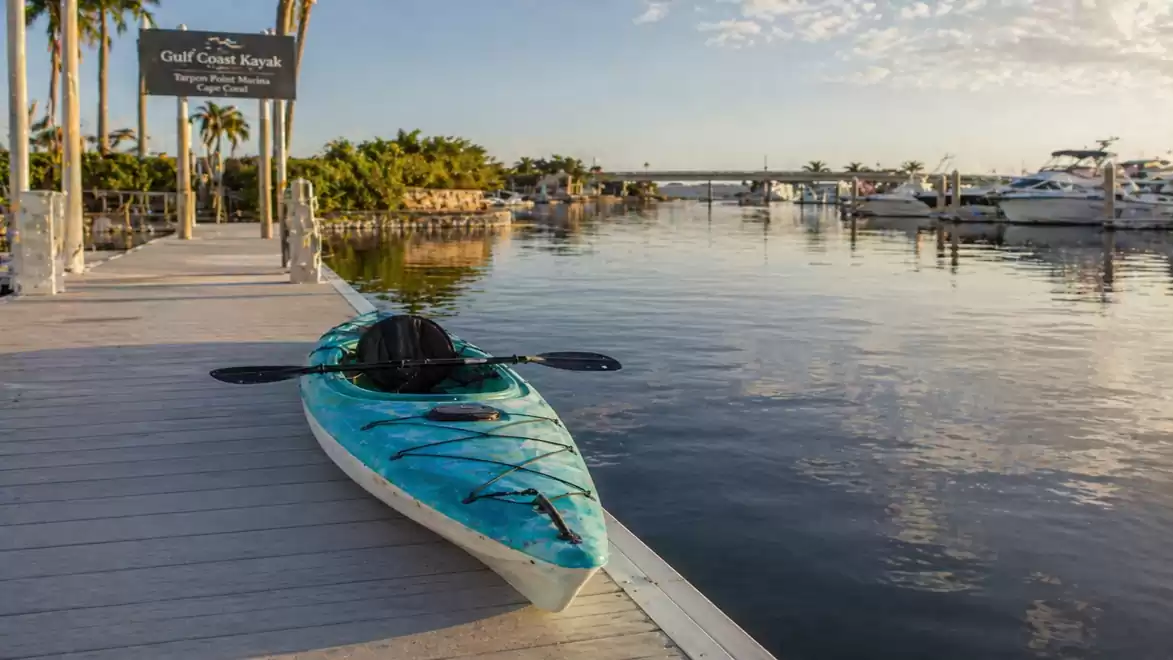 A blue kayak lies on a wooden dock in a marina with several yachts. The sky is partly cloudy and the sun is shining.