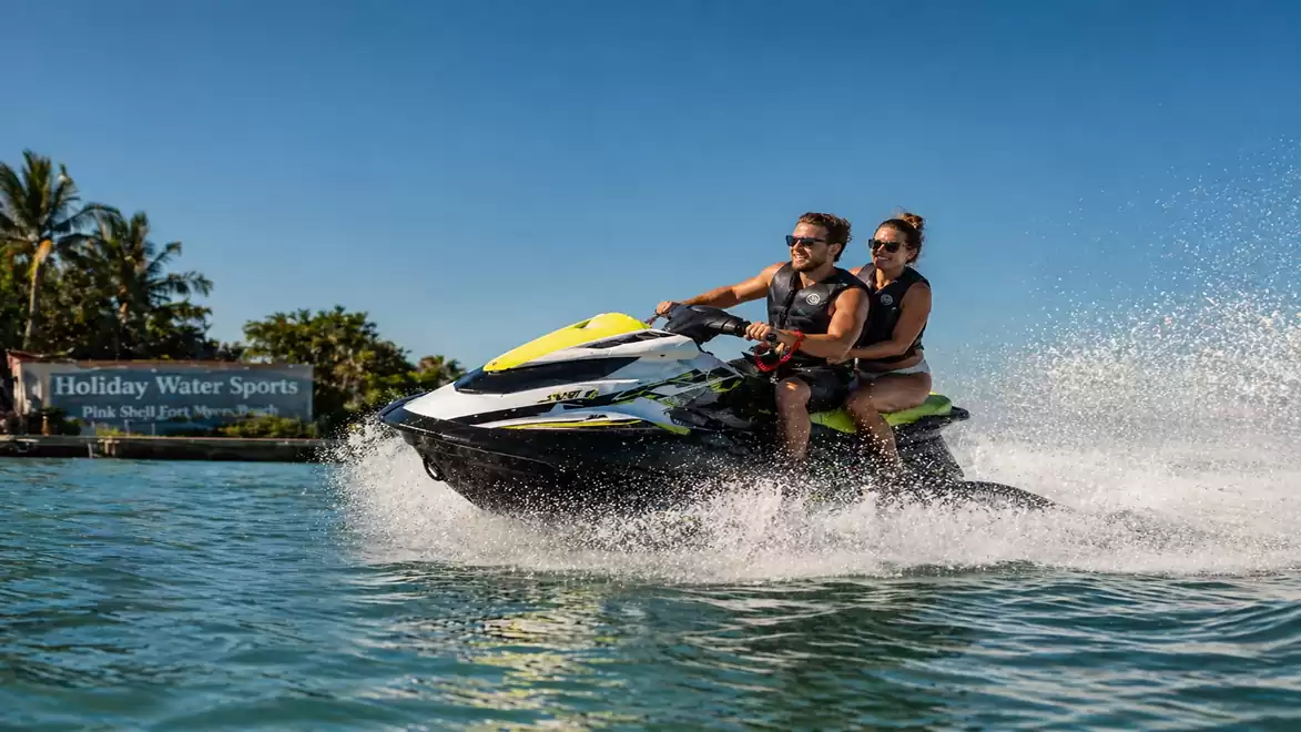 Two people riding a jet ski on the water in a sunny environment, surrounded by palm trees and blue sky.