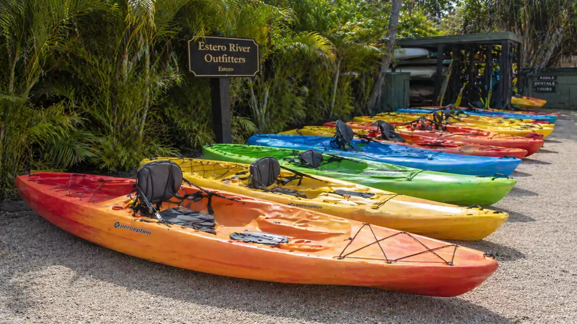 Colorful kayaks in various colors displayed on a gravel area at Estero River Outfitters, surrounded by green landscape.