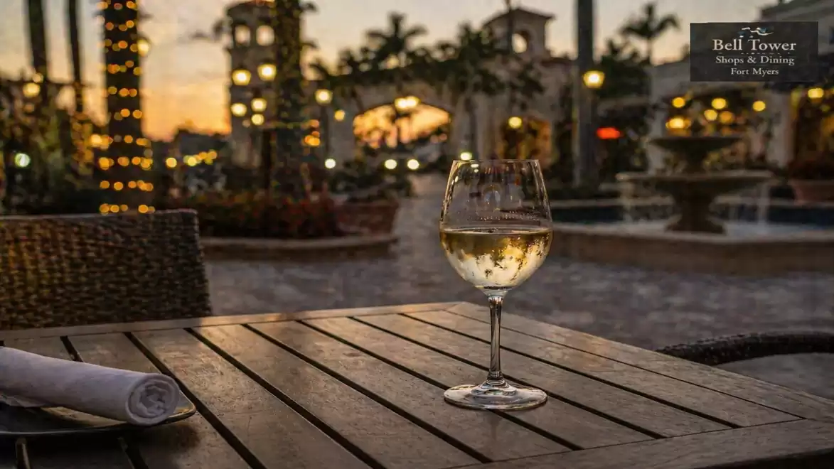 A glass of white wine on a wooden table in an outdoor setting, surrounded by festively lit buildings at sunset.