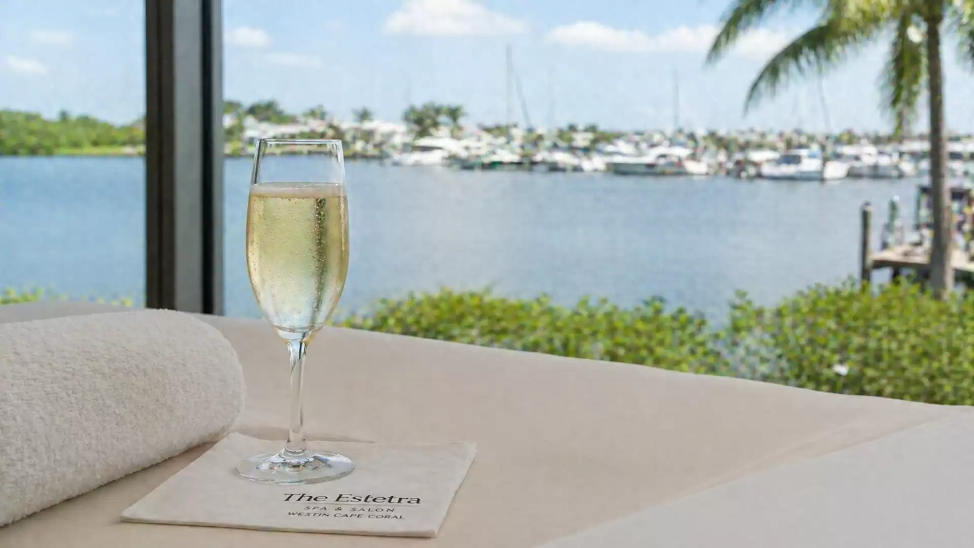 A glass of champagne on a table overlooking a tranquil harbor with sunny weather, surrounded by palm trees and yachts.