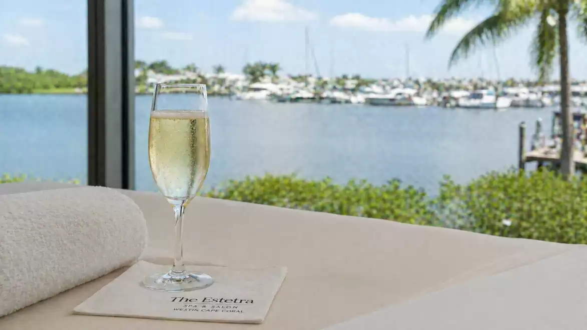 A glass of champagne on a table overlooking a tranquil harbor with sunny weather, surrounded by palm trees and yachts.