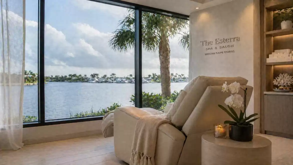 A luxurious spa room with a comfortable chair facing a tranquil lake view. A palm tree is visible in the background, and the light is softly dimmed to create a relaxing atmosphere.