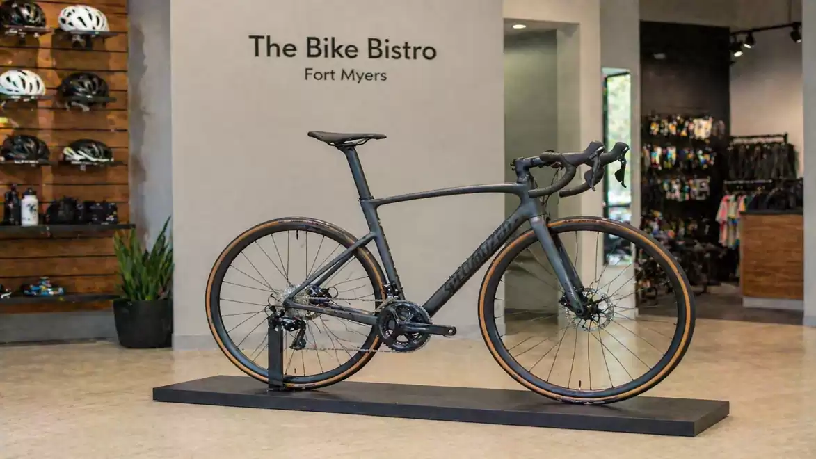 A modern road bike is displayed in a stylish bike shop called 'The Bike Bistro' in Fort Myers, surrounded by accessories and bicycle helmets on the wall.