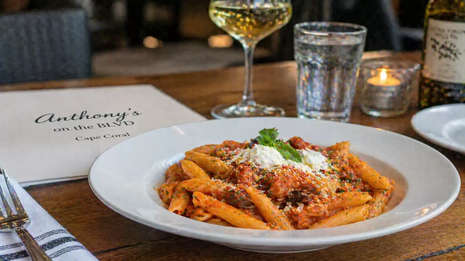 A plate of penne pasta in cream sauce, garnished with parsley and Parmesan. In the background, a glass of white wine and a candle.