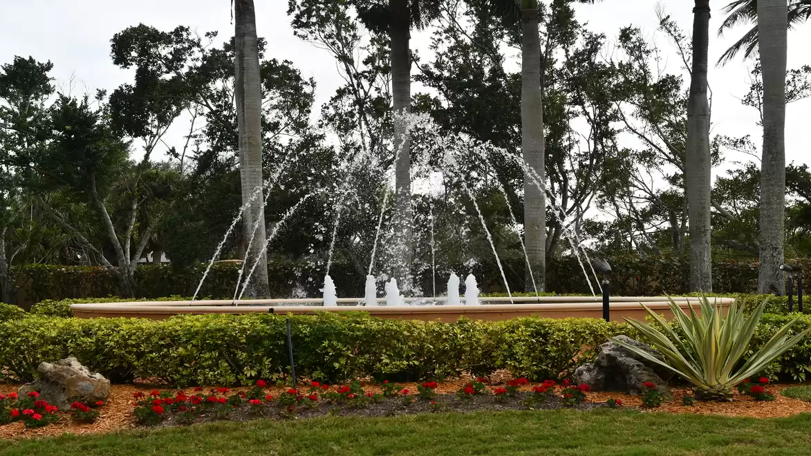 An elegant fountain in a garden surrounded by green bushes and colorful flowers. Tall trees rise in the background.