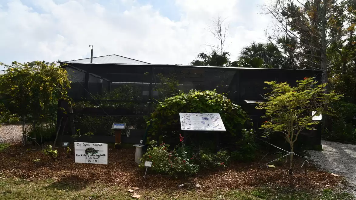 A large garden shed with black mesh, surrounded by green foliage and signs. The sky is cloudy.