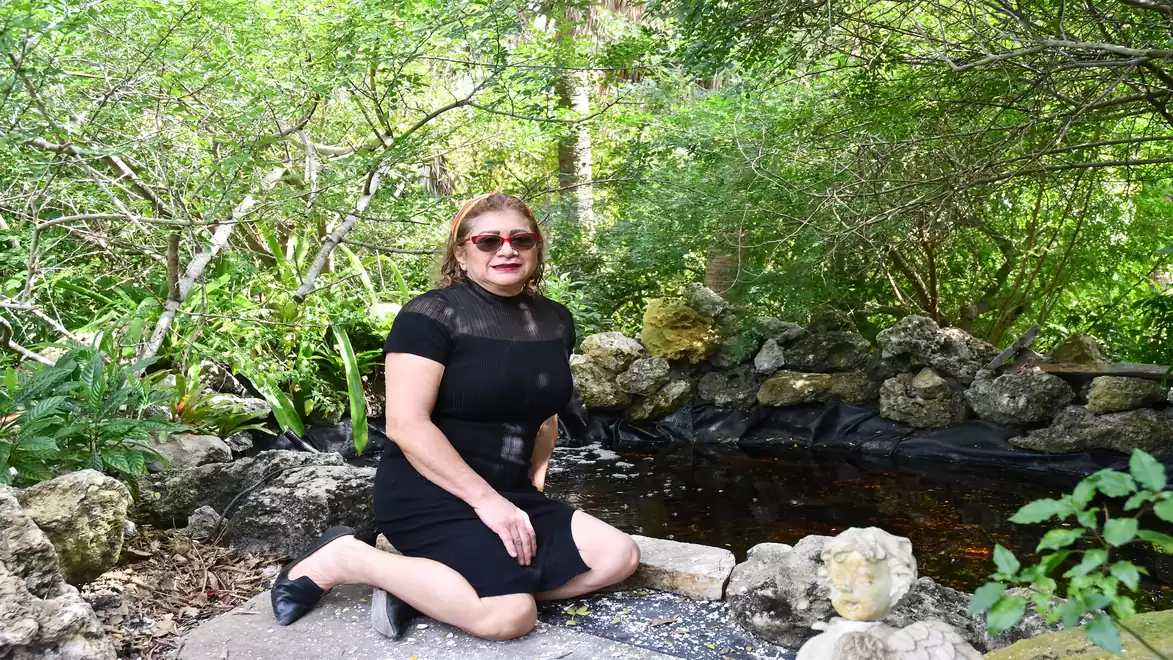 Woman in a black dress sits in front of a small pond in a lush, green garden.