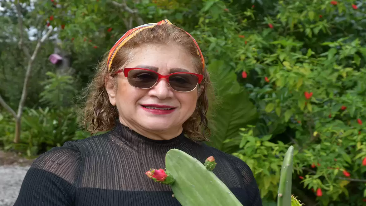 A woman wearing sunglasses and a floral headband stands in a garden surrounded by green plants.