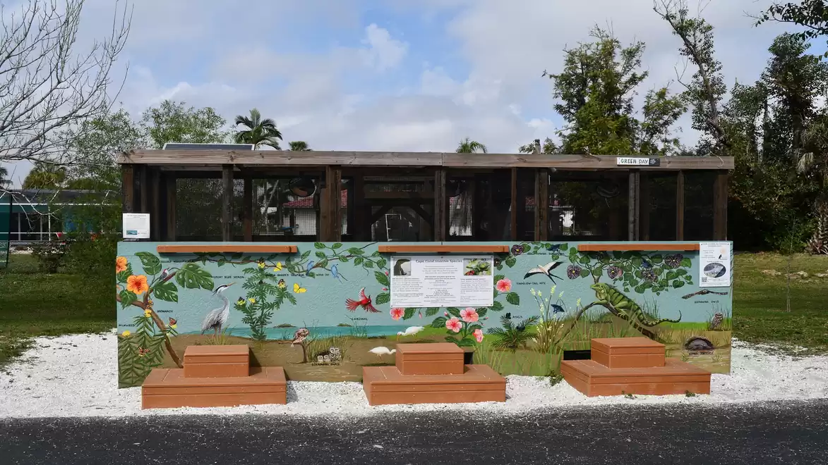 A wooden structure with a painted wall depicting a colorful array of wildlife and plants. In the foreground are steps and a gravel-covered ground.