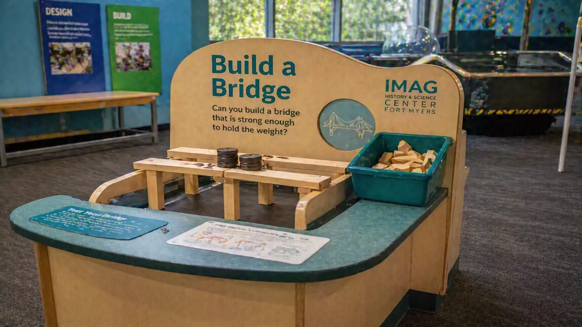 An interactive exhibit in a science center inviting people to build a strong bridge. The table has wooden blocks and a container with additional blocks.