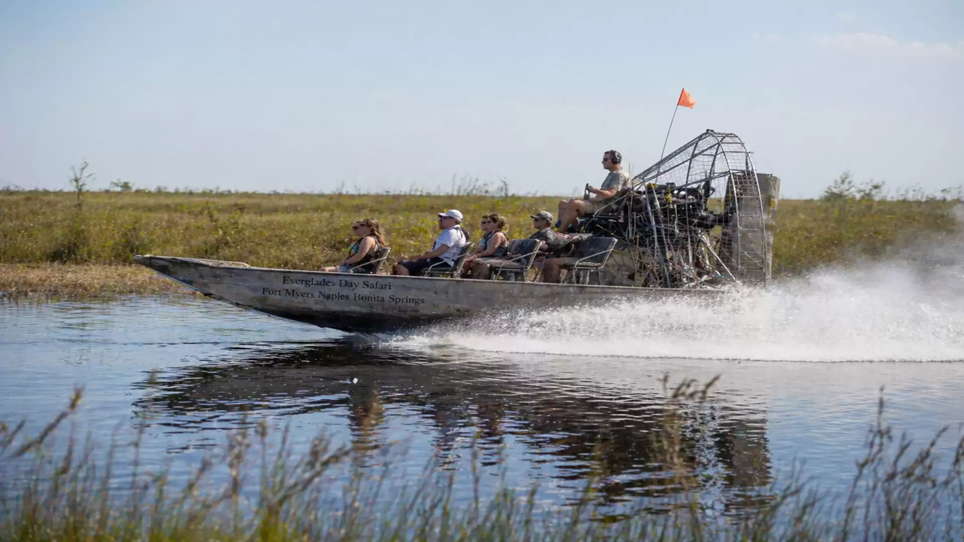An airboat with several people speeds across a shallow body of water in an open, grassy landscape.