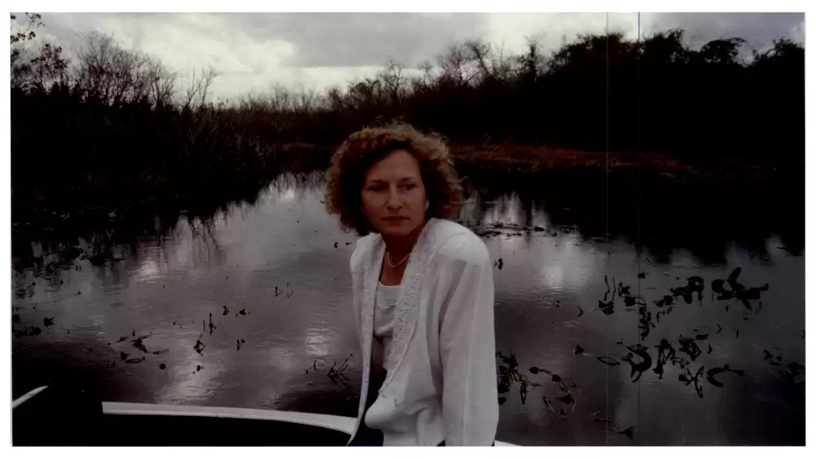 A person with curly hair sitting on a boat in front of a calm body of water, surrounded by natural scenery.