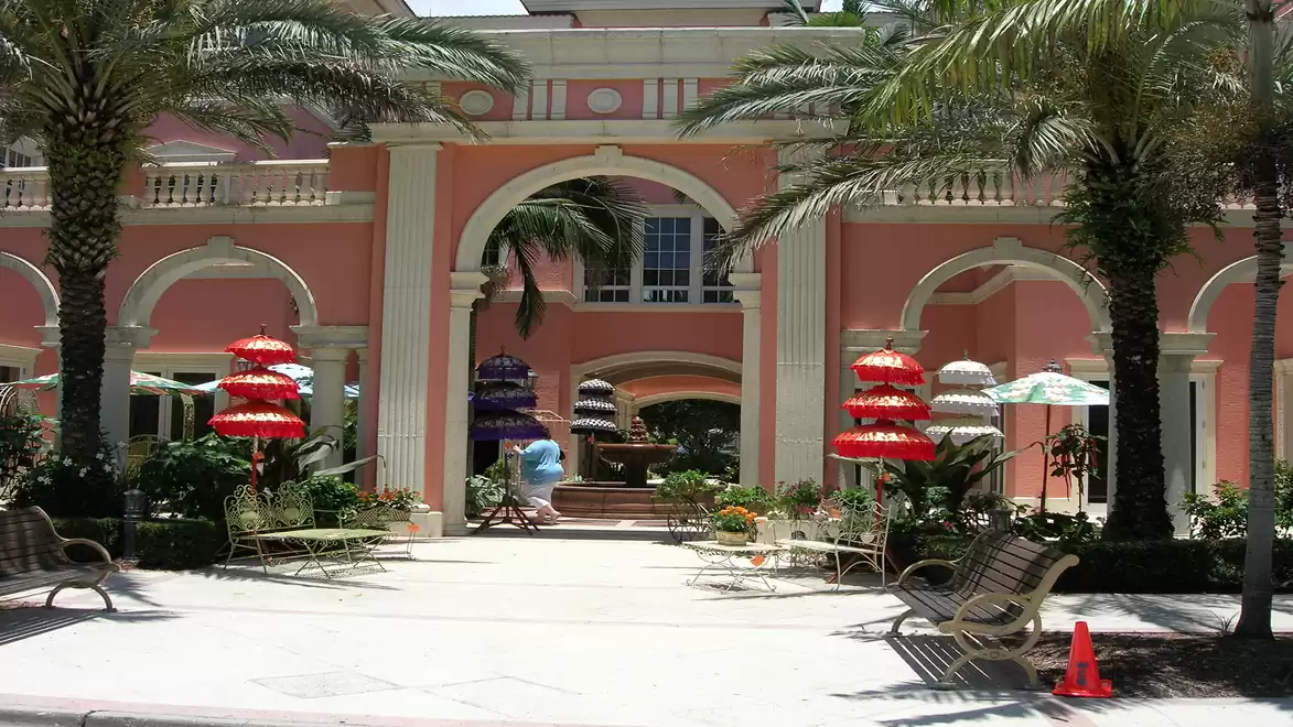 An elegant building with a pink facade, adorned with arches and palm trees. Colorful umbrellas and benches are visible in the foreground.