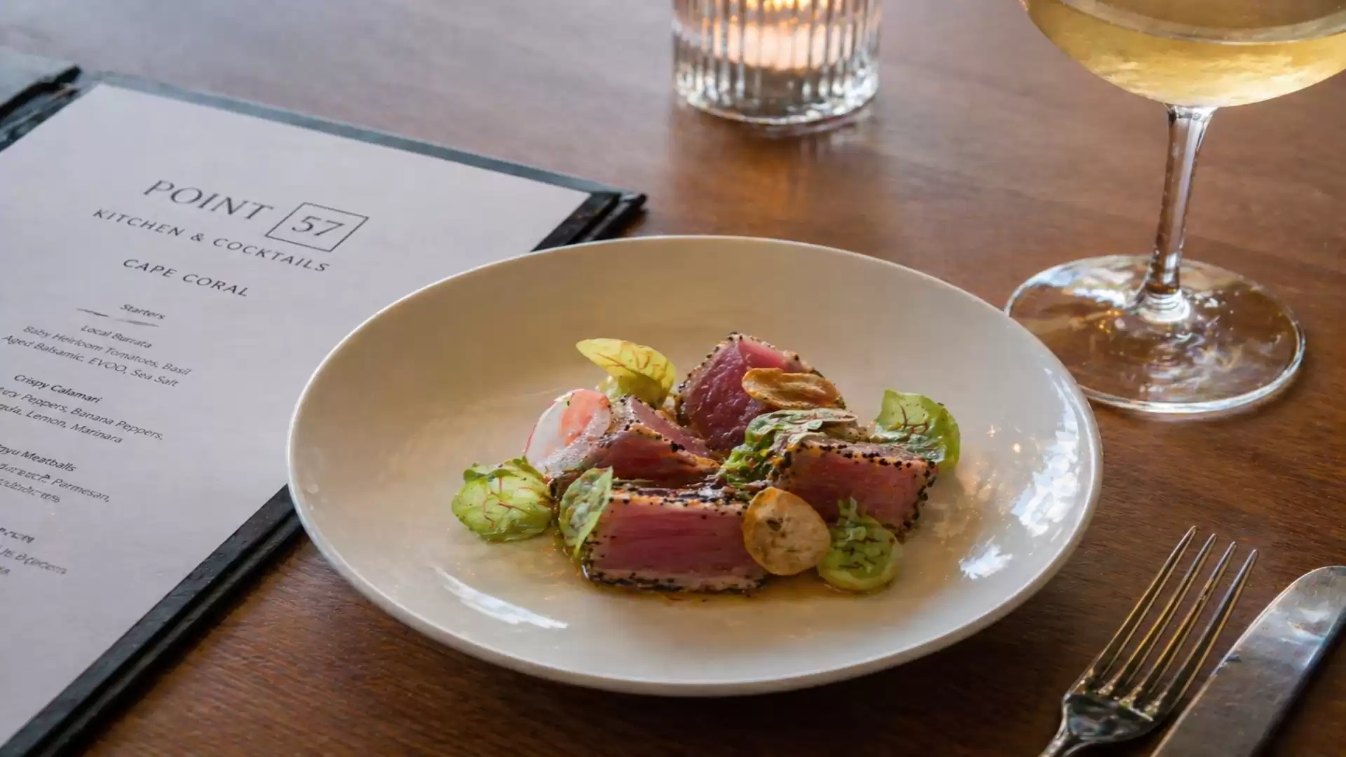 A stylishly set table featuring an elegant plate of tuna steak with sides, a glass of wine, a menu, and a lit candle.