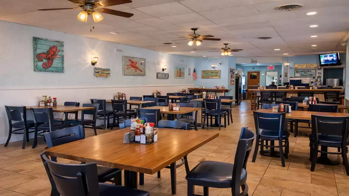 A cozy restaurant with wooden tables and blue chairs. Maritime decorations hang on the walls and fans are on the ceiling.