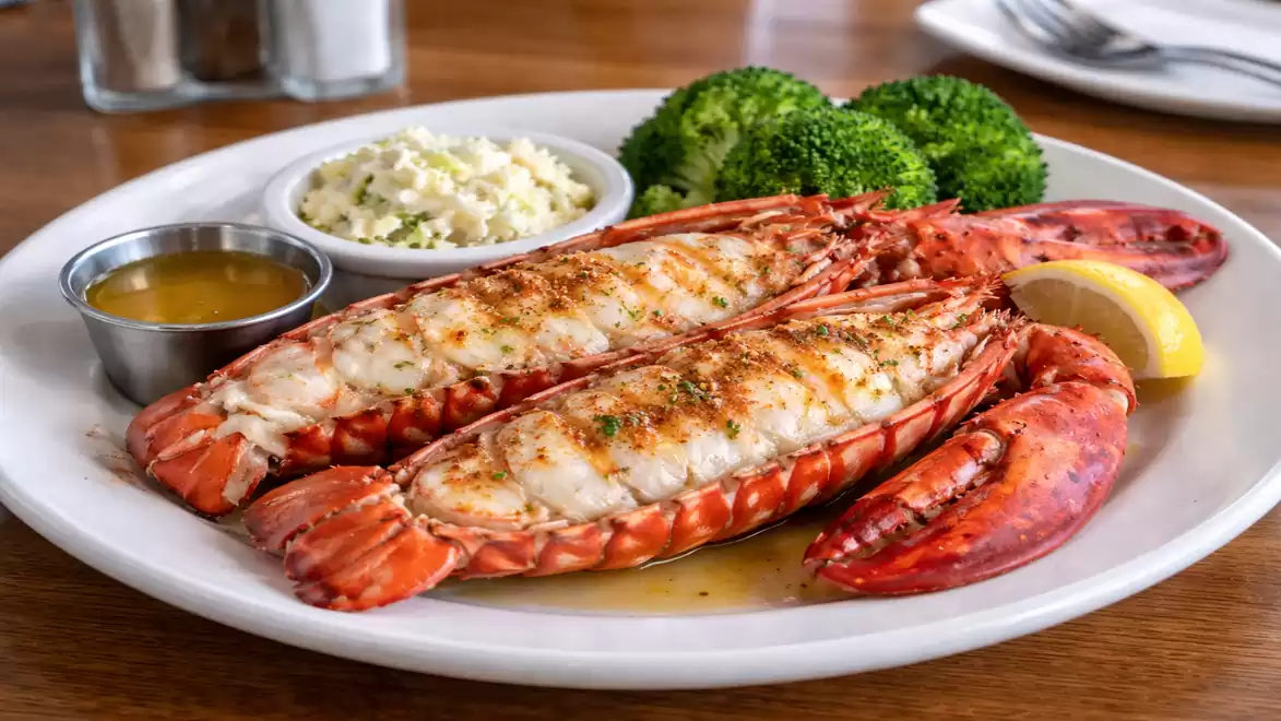 Grilled lobster on a white plate, served with broccoli, a dip in a metal cup, and a small bowl of coleslaw.