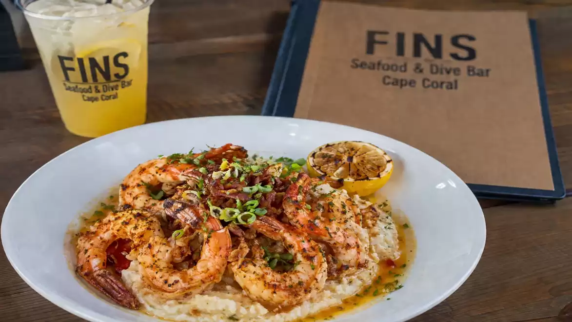 A plate of shrimp on risotto, garnished with herbs, next to a drink and a menu at FINS Seafood & Dive Bar in Cape Coral.
