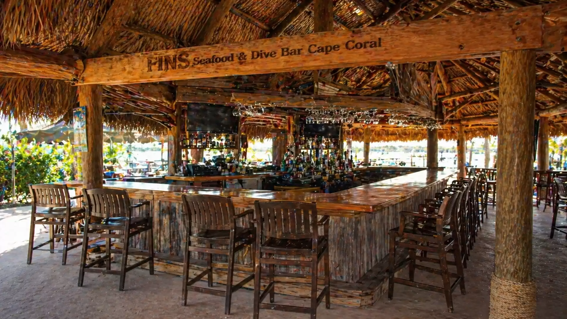 A tropical bar with a thatched roof and wooden furniture outdoors, many bar stools around the counter, with the sign 'PINS Seafood & Dive Bar Cape Coral' visible.