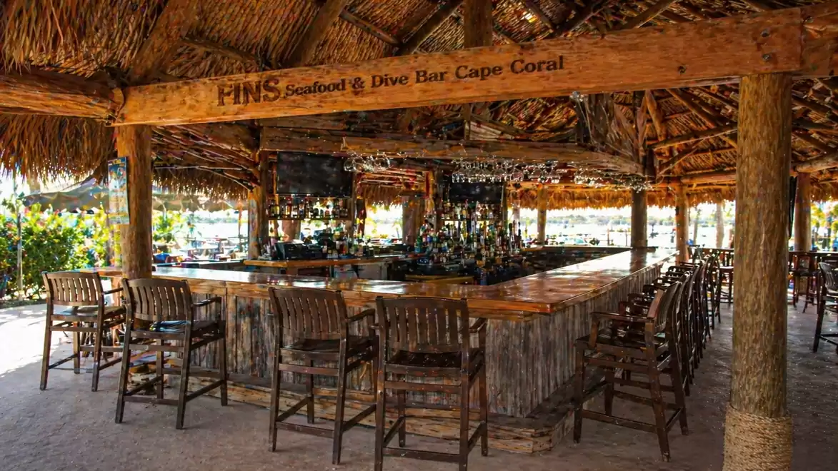 A tropical bar with a thatched roof and wooden furniture outdoors, many bar stools around the counter, with the sign 'PINS Seafood & Dive Bar Cape Coral' visible.