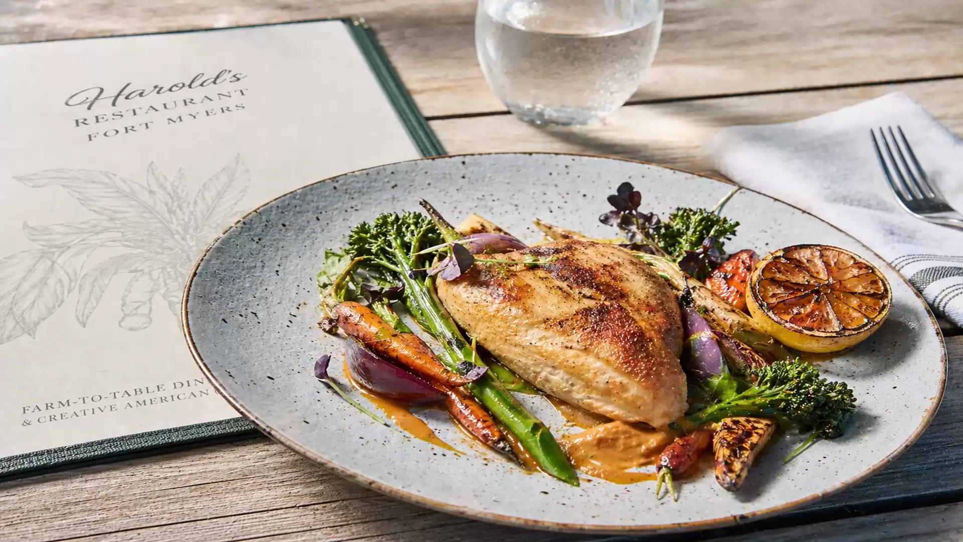 Elegant chicken dish with vegetables on a rustic wooden table, accompanied by a menu and a glass of water.